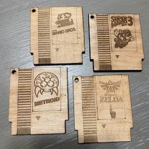 Peut inclure: Quatre ornements en bois de cartouche de jeu Nintendo, chacun représentant un jeu classique différent : Super Mario Bros. 3, Metroid, The Legend of Zelda et Super Mario Bros. Les ornements sont de couleur brun clair et ont une texture de grain de bois.