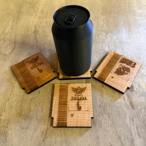 May include: A black can-shaped object sits on a wooden coaster. Three additional wooden coasters are visible, each designed to resemble classic video game cartridges. Two coasters feature "The Legend of Zelda" design, and one displays "Super Mario 3".
