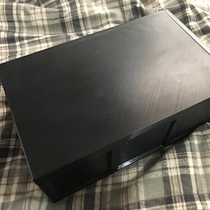 May include: A black rectangular box with a smooth, textured surface. The box has a slightly raised edge around the top and bottom.
