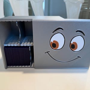 May include: A silver metal business card holder with a cartoon face design. The holder has a compartment for cards and a smiling face with large eyes. The cards are a dark blue color.
