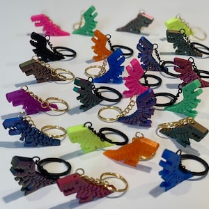 May include: A collection of 18 colorful dinosaur keychains. The keychains are made of plastic and feature a variety of colors, including red, blue, green, yellow, purple, and black. Each keychain has a metal ring for attaching to keys or bags.