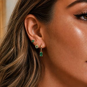 May include: Three gold earrings worn in a single ear. The top earring is a small green four-stone stud. The middle earring is a gold hoop with a flower-shaped charm featuring a central green stone. The bottom earring is a gold hoop with a green emerald-cut drop.