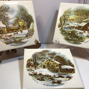 May include: Three ceramic tiles with winter landscape scenes. Each tile shows a different rural scene with buildings, trees, and snow. The tiles are displayed on decorative stands. The text on the tiles reads "The Farmer's Home Winter", "A Home in the Wilderness", and "The Old Homestead in Winter".