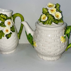 May include: White ceramic teapot and creamer set with a woven basket design. The teapot has a lid topped with white and yellow daisy flowers and green leaves. The handles are green and the creamer has a similar floral design.