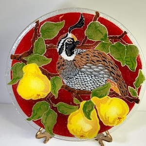 May include: A round glass plate with a red background and a painted design of a quail perched on a branch with pears. The quail is brown, gray, and white with black eyes. The pears are yellow with green leaves. The plate is on a gold stand.