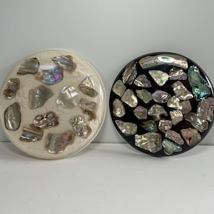 May include: Two round coasters. One is off-white with iridescent shell pieces, the other is black with similar shell pieces. The shell pieces have a pearlescent sheen, reflecting colors like green, blue, and purple.