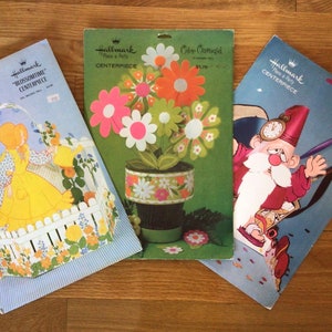May include: Three vintage Hallmark paper craft centrepieces. The first is a blue and white scene with a girl in a yellow dress holding a watering can. The second is a green and orange flower centrepiece. The third is a blue and white scene with a gnome holding a clock.