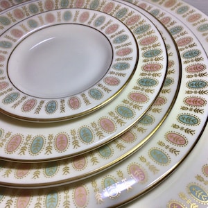 May include: A set of vintage white ceramic plates with a gold rim and a repeating pattern of pink, blue, and gold oval shapes. The plates are stacked, showcasing different sizes, from a small saucer to a large dinner plate.