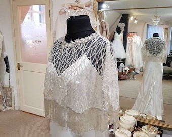 Puede incluir: Un vestido de novia blanco con una superposición de cuentas y flecos en un maniquí. El vestido se exhibe en una tienda de novias con un espejo que refleja otros vestidos. El texto "Breath Away" es visible en la pared.