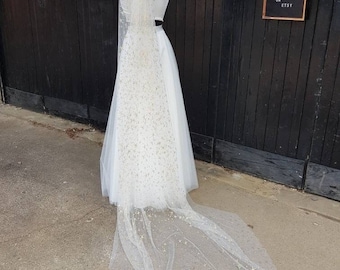 May include: A white wedding dress and veil on a mannequin. The veil is long and sheer, adorned with small star-shaped embellishments. The dress is simple and elegant, with a fitted bodice and flowing skirt. A sign on the right reads "Bexbrides Boho Bespoke Quirky Vintage Eclectic Brides on Etsy."
