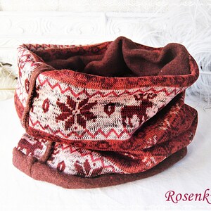 Könnte beinhalten: Ein dunkelroter und cremefarbener Schlauchschal mit einem wiederkehrenden Muster aus Rentieren und Schneeflocken. Der Stoff hat eine strukturierte Strickaußenseite und ein einfarbig dunkelrotes Fleecefutter. Der Text Rosenknopf steht unten rechts.