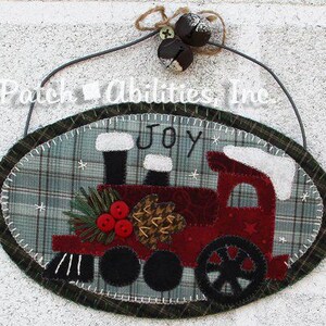May include: An oval fabric wall hanging featuring a red train applique on a green plaid background with the text "JOY" above it. The train is decorated with three red buttons, pine needles, and a pinecone. Two dark metal bells hang from a wire.