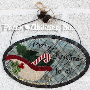 May include: An oval fabric wall hanging featuring a plaid background with a stitched applique of a sleigh carrying a green tree and a candy cane. The text Merry Christmas to all is embroidered in black. It includes a wire hanger with two jingle bells.