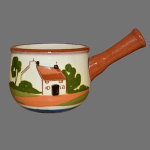 May include: A small ceramic soup bowl with a handle. The bowl features a painted cottage scene in white, orange, green, and brown. The handle is a solid orange-brown, and the rim is a darker brown. A charming kitchenware item.