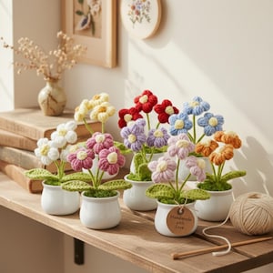 May include: A collection of handmade crochet flowers in small white pots. The flowers are in various colors, including white, yellow, red, purple, blue, pink, and orange. Each pot has green leaves and a brown stem. A tag reads "Handmade with Love."