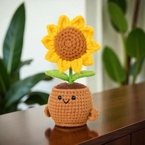 May include: A crocheted sunflower in a brown pot with a smiling face. The sunflower has yellow petals and a brown center. The pot has small arms. The craft is likely handmade.