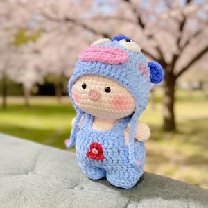 May include: A handmade crochet doll wearing a light blue outfit and a matching hat. The doll has a light skin tone, rosy cheeks, and black eyes. The hat features pink and blue accents. A red octopus is sewn on the front of the overalls. The doll is sitting on a gray surface.