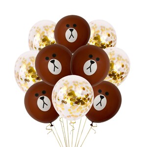 Pu&ograve; includere: Un mazzo di palloncini marroni con un viso di orso disegnato su di essi. I palloncini sono legati insieme con uno spago dorato. Alcuni dei palloncini sono trasparenti con coriandoli dorati all'interno.