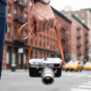 Op de afbeelding: Een zilveren en zwarte Leica M-serie camera met een bruine leren riem wordt voor een stadsstraat gehouden. De cameralens is scherp en de riem is aan de camerabehuizing bevestigd.