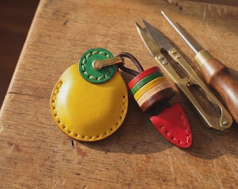 Op de afbeelding: Een rond geel lederen tasje met een groen cirkelvormig accent, bevestigd aan een veelkleurige lederen bedel met gestapelde rode, groene, gele en bruine lagen, eindigend in een rode punt, naast een metalen schaar en een priem.