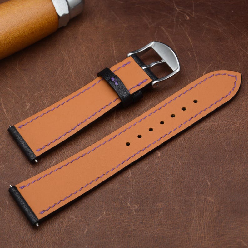 May include: A two-piece watch strap featuring a tan leather interior with purple stitching and black leather edges. The strap includes a silver-toned metal buckle and a black leather keeper loop, with spring bars visible at the attachment ends.