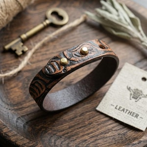 Pode incluir: Uma pulseira de couro castanho com um design entalhado e tachas de latão. A pulseira está numa bandeja de madeira com uma chave antiga e uma etiqueta que diz "- LEATHER -."