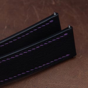 May include: A black watch strap featuring a textured surface and vibrant purple stitching along the edges. The strap includes integrated metal spring bars at the top ends for attachment, showcasing a clean design with contrasting thread colors.