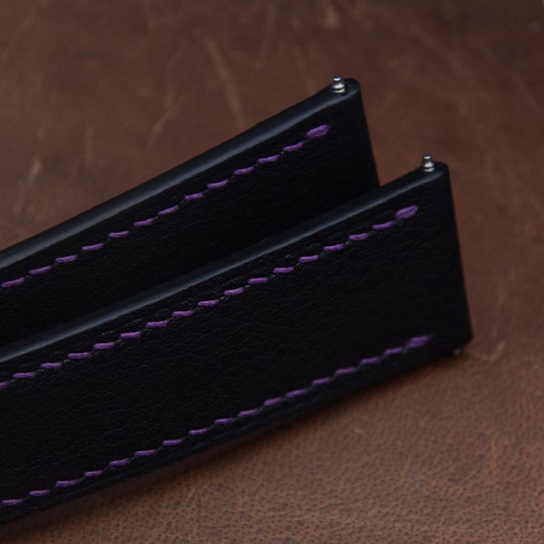 May include: A black watch strap featuring a textured surface and vibrant purple stitching along the edges. The strap includes integrated metal spring bars at the top ends for attachment, showcasing a clean design with contrasting thread colors.