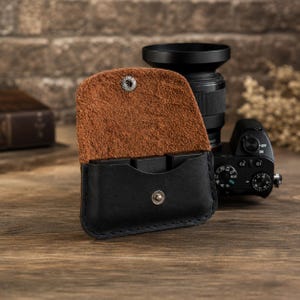 May include: Black leather camera memory card holder with a brown suede interior. The holder is open, revealing the card slots. A silver snap closure secures the flap. A camera and lens are in the background.