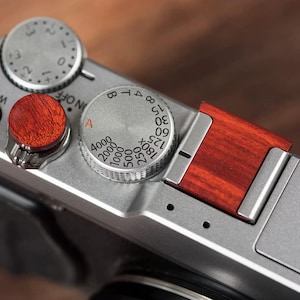 May include: Close-up of a silver digital camera with wooden accents. The camera features a red wooden shutter button and a wooden grip. The dials are marked with numbers and letters, indicating settings for aperture and shutter speed.