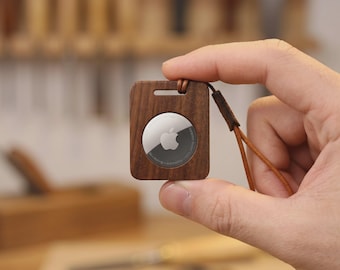 Op de afbeelding: Een vierkante houten AirTag-houder met een bruin leren riempje. De houder heeft een uitsparing voor de AirTag, die een zilveren oppervlak met het Apple-logo heeft. De houder wordt in een hand gehouden, met een vage werkplaatsachtergrond.