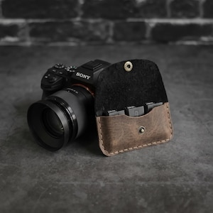 May include: A brown leather memory card holder with a snap closure, open to reveal several memory cards. A black Sony camera with a lens is in the background. The holder has a dark interior and is stitched around the edges.