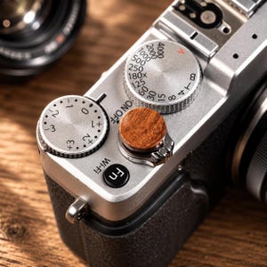 Peut inclure: Le dessus d'un appareil photo numérique argenté avec un déclencheur circulaire en bois, une molette de compensation d'exposition argentée graduée de -3 à +3, une molette de vitesse d'obturation et un bouton noir marqué Fn et Wi-Fi.