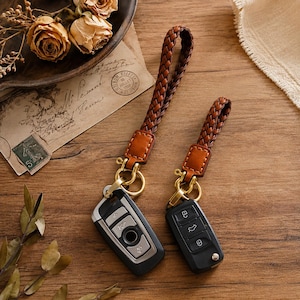 May include: Two keychains with braided brown leather straps and gold-colored hardware. One keychain is attached to a black car key fob, the other to a silver and black key fob. The keychains rest on a wooden surface.