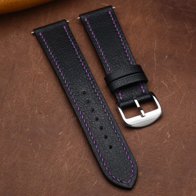 May include: A black leather watch strap featuring a textured surface and vibrant purple stitching along the edges. The strap includes a polished silver-tone metal buckle and two matching leather keepers, designed for secure attachment to a timepiece.