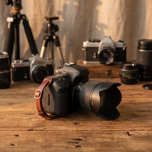 May include: A collection of photography equipment, including a black DSLR camera with a brown leather strap, lenses, and tripods, arranged on a wooden surface. Several vintage film cameras are also visible, suggesting a photography theme.