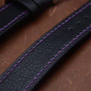 May include: A black watch strap with a textured surface and vibrant purple stitching running along both edges. The strap features a series of pre-punched holes for adjustment and a matching black leather loop to secure the end of the band.