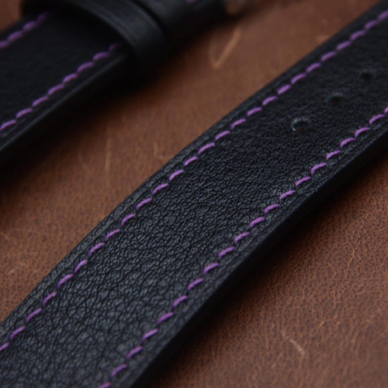 May include: A black watch strap with a textured surface and vibrant purple stitching running along both edges. The strap features a series of pre-punched holes for adjustment and a matching black leather loop to secure the end of the band.