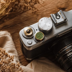 May include: A silver camera top plate featuring a circular brass dial, a textured green leather shutter release button, and a metallic silver exposure dial with engraved letters and numbers, including a hot shoe mount on the right side.