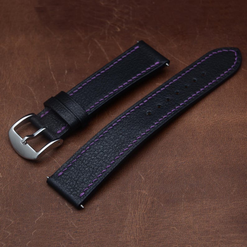May include: A black leather watch strap featuring purple contrast stitching. The two-piece band includes a silver-toned metal buckle and two leather keepers. The textured surface of the leather is visible along the length of both strap pieces.