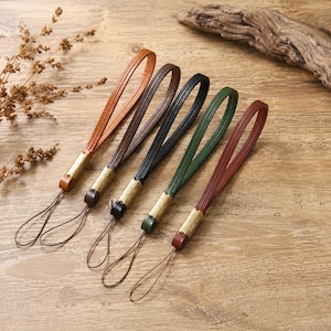 May include: Five leather wrist straps in various colors: tan, brown, black, green, and burgundy. Each strap features a loop for the wrist and a small loop for attaching to a phone or other device. The straps have gold-colored metal accents.