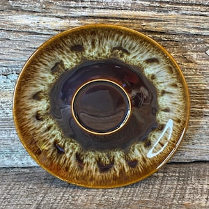 May include: A vintage ceramic saucer with a brown and cream glaze. The saucer has a deep brown center with a gold rim and a textured, speckled pattern around the edges. The overall design is reminiscent of mid-century modern pottery.