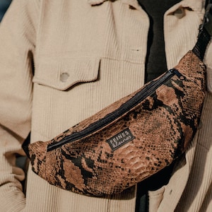 Op de afbeelding: Een bruine heuptas met een slangenhuidpatroon, een zwarte band en de tekst "LEINES" in witte letters. De heuptas wordt gedragen door iemand die een beige corduroy jack draagt.