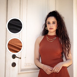 Könnte beinhalten: Eine Frau trägt ein rostiges, geripptes Tanktop und eine passende Hose. Das Oberteil hat einen Stehkragen und die Hose ist weit geschnitten. Sie steht vor einer weißen Tür mit einer goldenen Kettenkette.