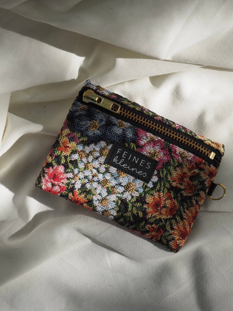 May include: A small black zippered pouch with a floral tapestry design. The pouch has a gold zipper and a black label that reads "FEINES kleines".
