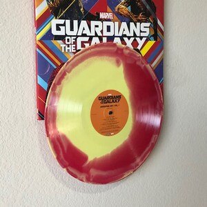 May include: A vinyl record with a yellow and red swirl design, featuring the text "Awesome Mix Vol. 1". The record is displayed in front of a Marvel "Guardians of the Galaxy" poster with character illustrations and the movie title.