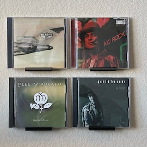 May include: Four CD cases on a white wall. The cases are black with clear plastic fronts. The CD covers feature the following artists: Beastie Boys, Kid Rock, Fleetwood Mac, and Garth Brooks.