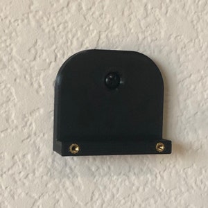 May include: A black, 3D-printed bracket with a rounded top and a small shelf. The bracket is mounted on a textured, off-white wall. Two brass-colored inserts are visible on the shelf, and a screw secures the bracket to the wall.