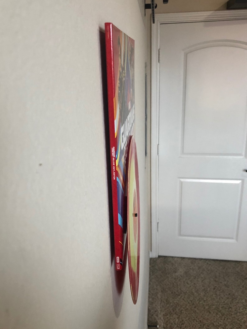 May include: A red and yellow vinyl record mounted on a wall, with a red rectangular album cover behind it. The album cover has text and images, and the record is circular with a yellow center.