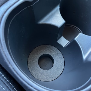 May include: A black car cup holder with a textured, circular insert. The insert has a hole in the center and sits inside the cup holder. The cup holder is part of a car's interior.